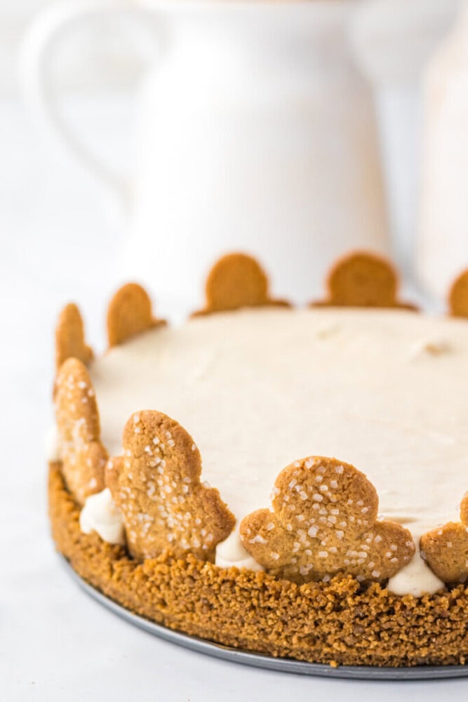A cheesecake with a graham cracker crust, topped with frosting and decorated around the edge with gingerbread cookies, sits on a white surface.