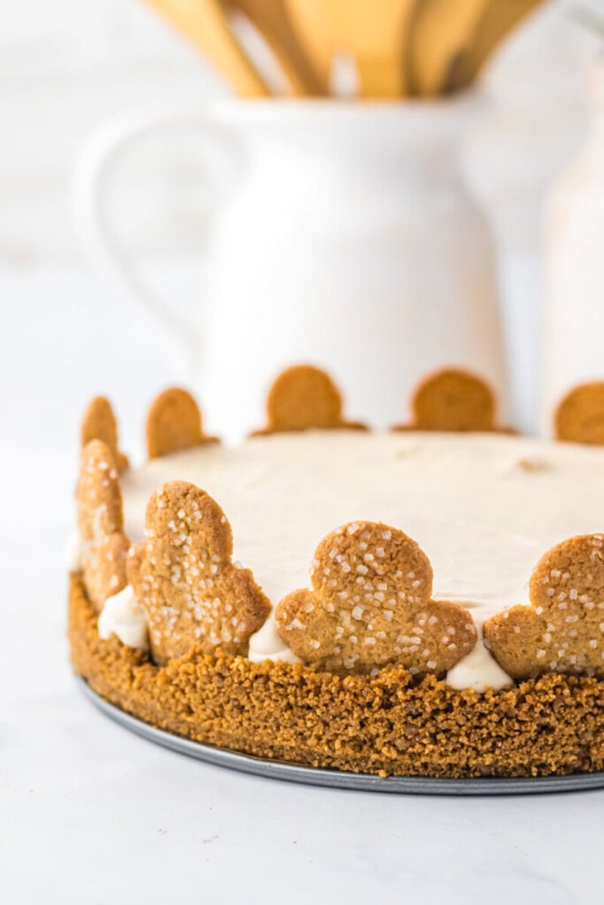 A cheesecake with a crumb crust, topped with gingerbread cookies arranged upright around the edge, sits on a white surface with a white pitcher in the background.