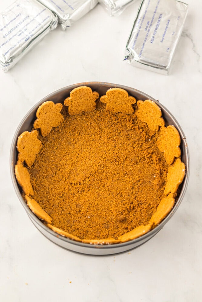 A round baking pan with a crumb crust bottom and gingerbread cookies lining the sides; unopened cream cheese blocks are in the background.