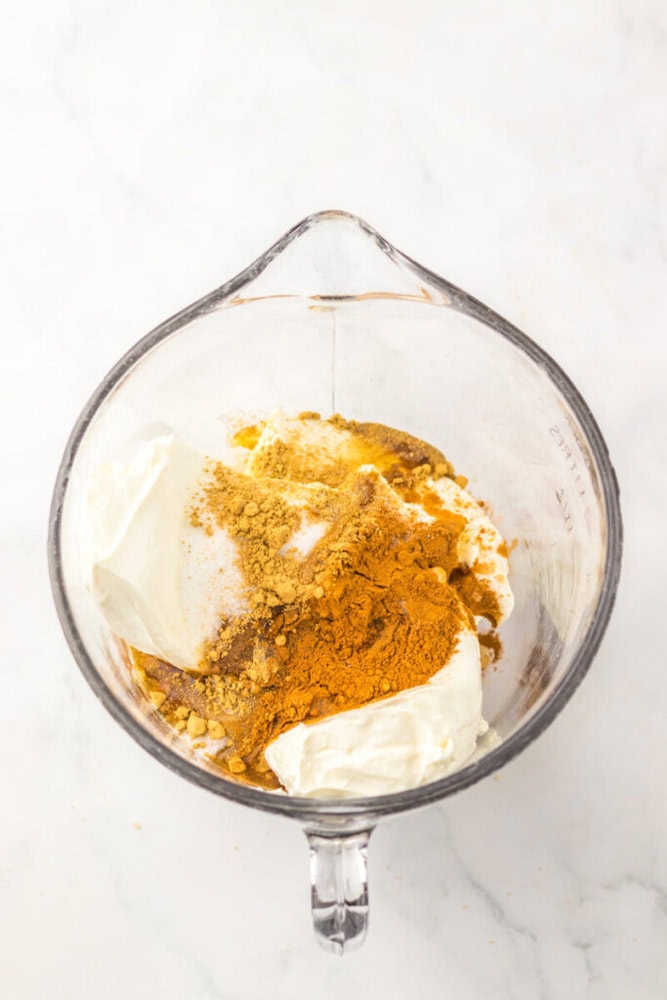 Glass mixing bowl containing yogurt, cream cheese, and various yellow and brown powdered spices on a white marble surface.