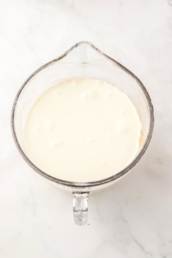 A clear glass measuring cup filled with a white liquid, likely cream or milk, sits on a white marble surface.