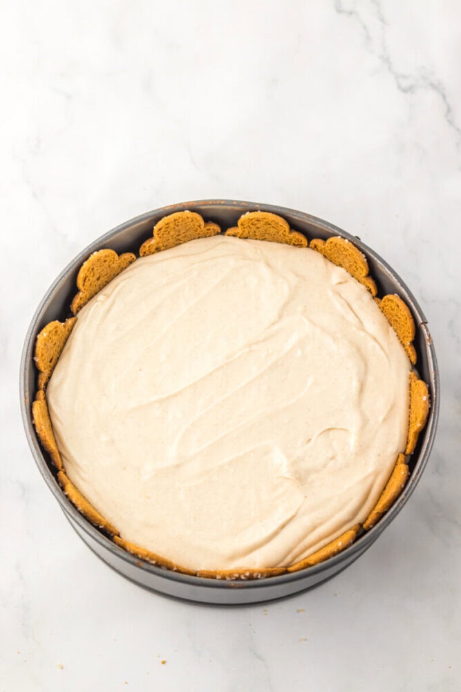 A round cheesecake with a smooth filling, lined with gingerbread cookies around the edge, in a springform pan on a white marble surface.