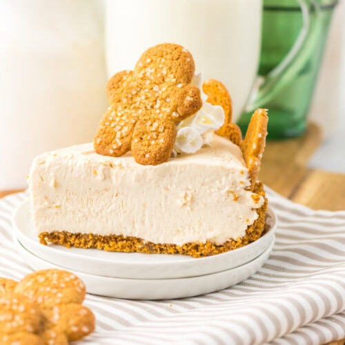 A slice of cheesecake with a graham cracker crust, topped with whipped cream and a gingerbread cookie, served on a white plate.