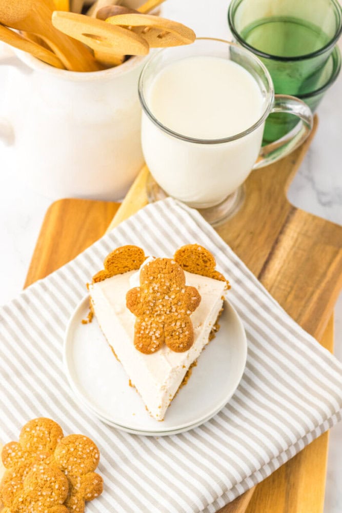 A slice of cake topped with a gingerbread cookie on a plate, beside a glass of milk, wooden utensils, and stacked cups on a wooden tray.