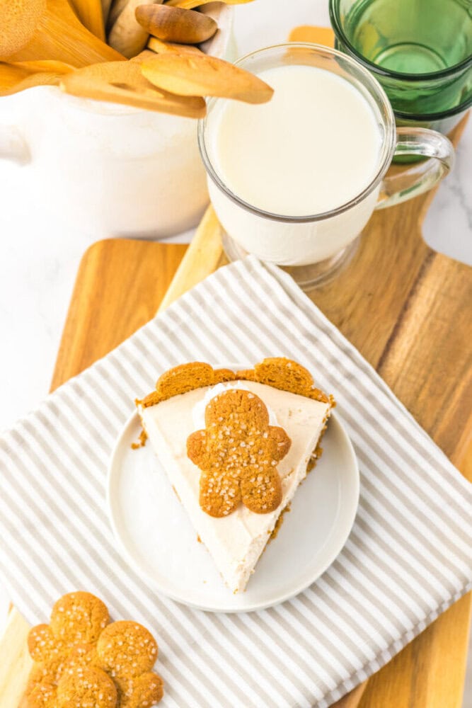 A slice of pie topped with a gingerbread cookie on a white plate, next to a glass of milk and a wooden container holding utensils.