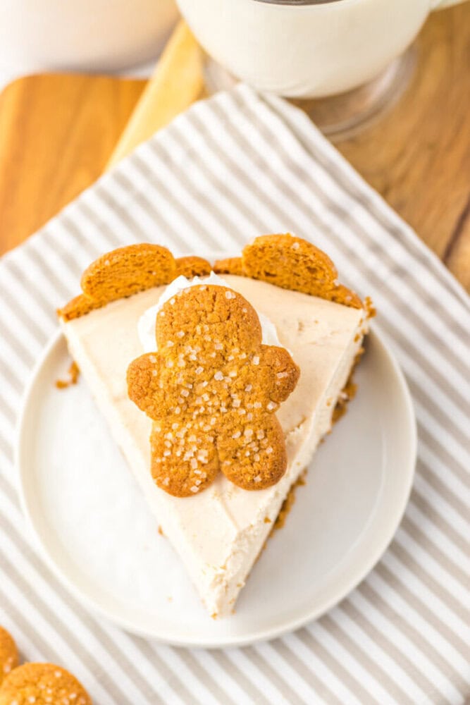 A slice of cheesecake topped with whipped cream and a gingerbread cookie shaped like a person, served on a white plate with a striped napkin underneath.