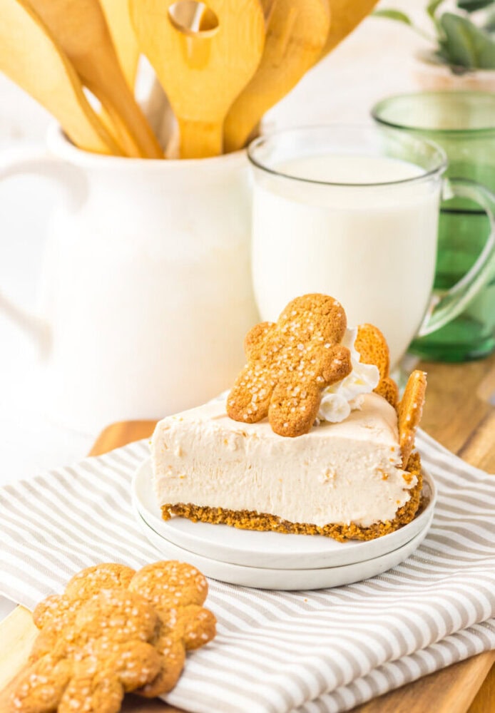 A slice of cheesecake topped with whipped cream and a gingerbread cookie sits on a plate, with more cookies, a glass of milk, and kitchen utensils in the background.