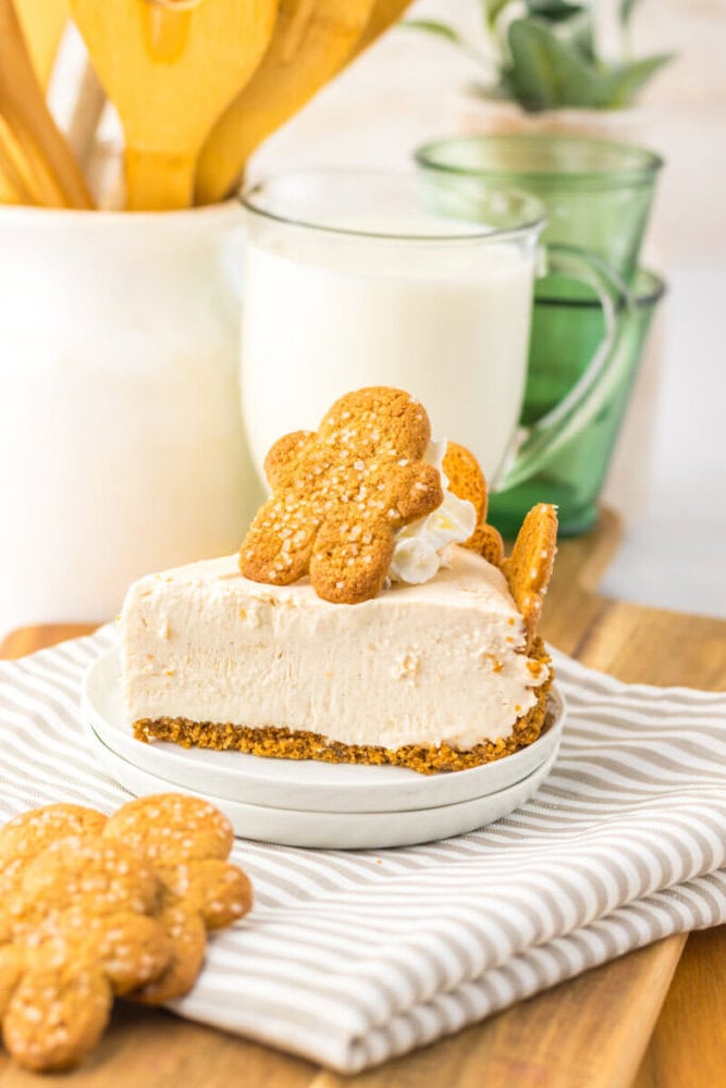 A slice of creamy pie with a graham cracker crust, topped with whipped cream and gingerbread cookies, served on a white plate with a glass of milk in the background.