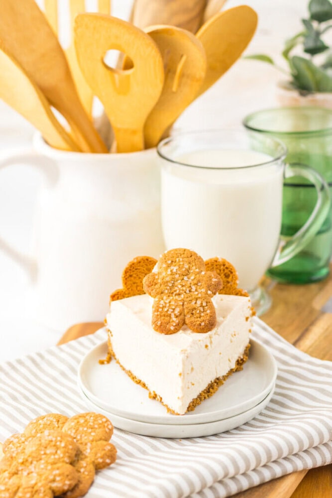 A slice of cheesecake with a gingerbread cookie on top sits on a plate next to cookies, a glass of milk, and a white pitcher holding wooden utensils.