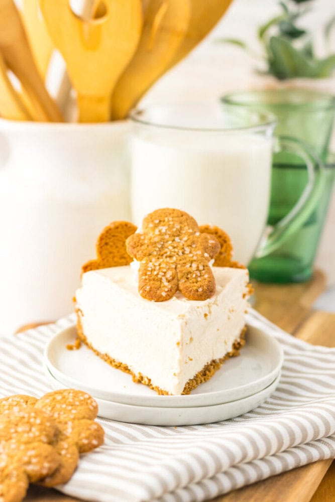A slice of cheesecake with a gingerbread cookie on top sits on a white plate, with a glass of milk and kitchen utensils in the background.