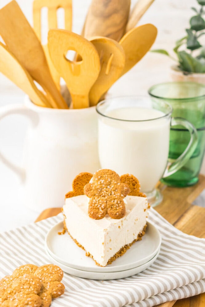 A slice of cheesecake topped with gingerbread cookies is on a plate next to a glass mug of milk, wooden utensils in a white holder, and two green glasses in the background.