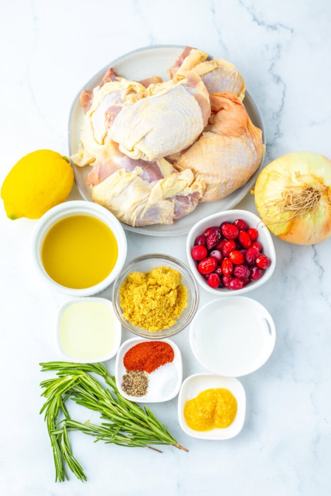 Raw chicken pieces on a plate surrounded by a lemon, onion, cranberries, olive oil, brown sugar, white vinegar, spices, garlic paste, and fresh rosemary on a white surface.