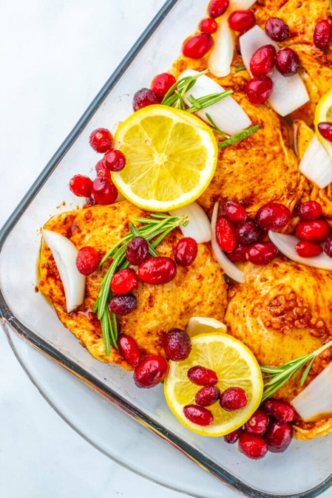 A glass baking dish containing seasoned chicken thighs topped with lemon slices, fresh cranberries, onion chunks, and rosemary sprigs.