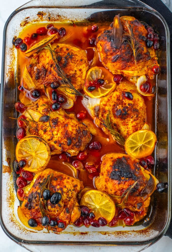 Baked chicken thighs in a glass dish, garnished with lemon slices, cranberries, olives, rosemary, and onions in a seasoned sauce.
