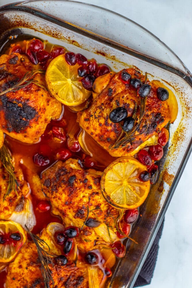 Baked chicken thighs in a glass dish with lemon slices, cranberries, rosemary sprigs, and a red seasoned sauce.