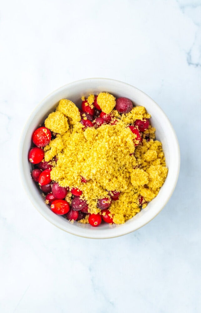 A white bowl filled with fresh cranberries topped with a mound of brown sugar, set on a light marble surface.