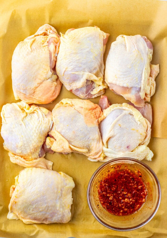 Six raw chicken thighs on parchment paper next to a glass bowl of red marinade or sauce.
