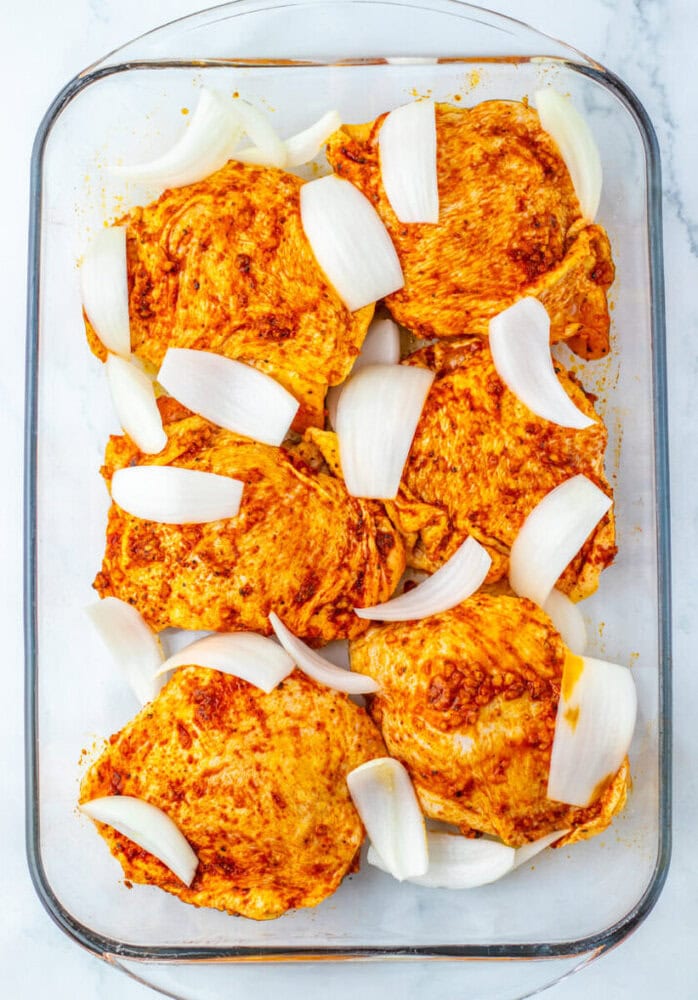 Raw, seasoned chicken thighs and large onion pieces arranged in a glass baking dish on a light surface.