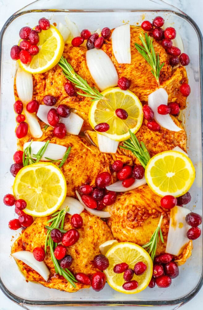 Raw seasoned chicken thighs in a glass baking dish, topped with lemon slices, cranberries, rosemary sprigs, and onion chunks, ready to be cooked.