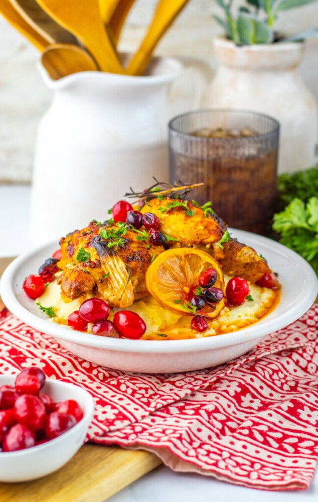 A plate of roasted chicken with lemon, cranberries, and herbs served over mashed potatoes, with a small bowl of cranberries and a glass of soda in the background.