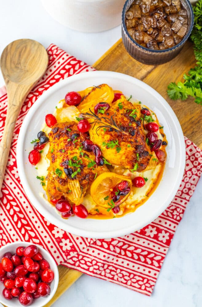 A plate of roasted chicken breasts with lemon slices, cranberries, and herbs on mashed potatoes, next to a bowl of fresh cranberries and a jar of roasted mushrooms.