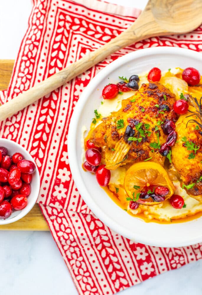 Baked chicken with lemon slices and cranberries on a white plate, garnished with herbs, next to a bowl of fresh cranberries on a red patterned cloth.
