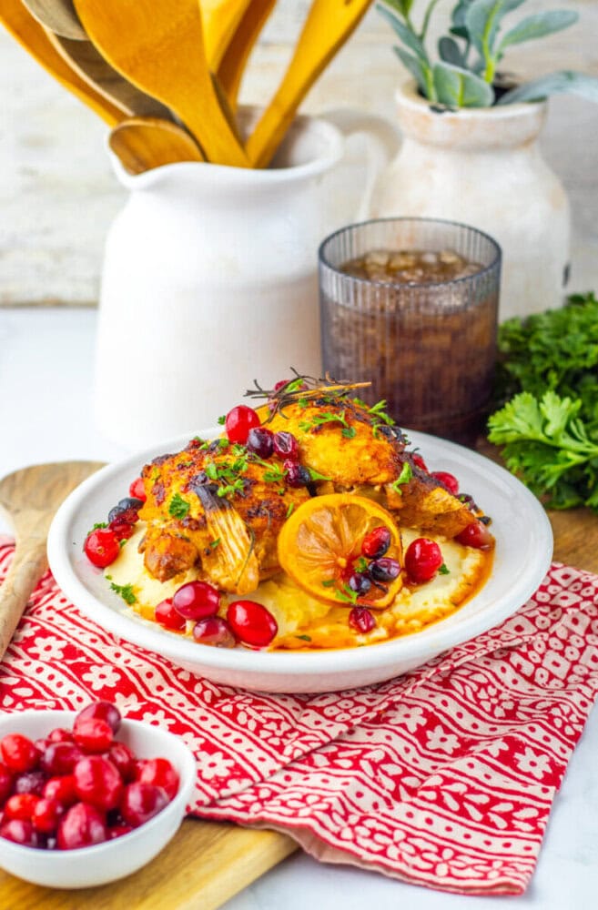 A plate of roasted chicken with lemon, cranberries, and herbs served on mashed potatoes, garnished with fresh parsley, next to a glass of drink and a bowl of cranberries.
