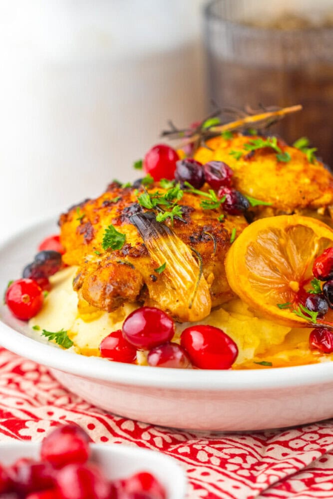 Roast chicken served over mashed potatoes, garnished with lemon slices, fresh cranberries, and chopped herbs on a white plate.