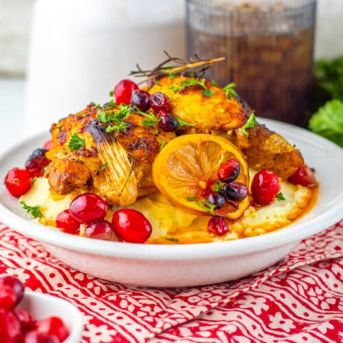 A plate of roasted chicken garnished with lemon, cranberries, and parsley, served over mashed potatoes on a patterned red cloth.