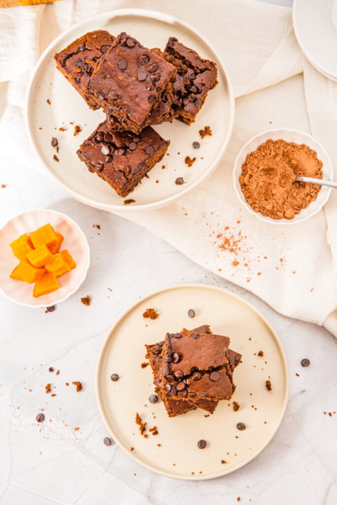 Two plates with chocolate chip brownies, a small bowl of cocoa powder, and a dish with cubed orange fruit on a white surface with scattered chocolate chips and crumbs.