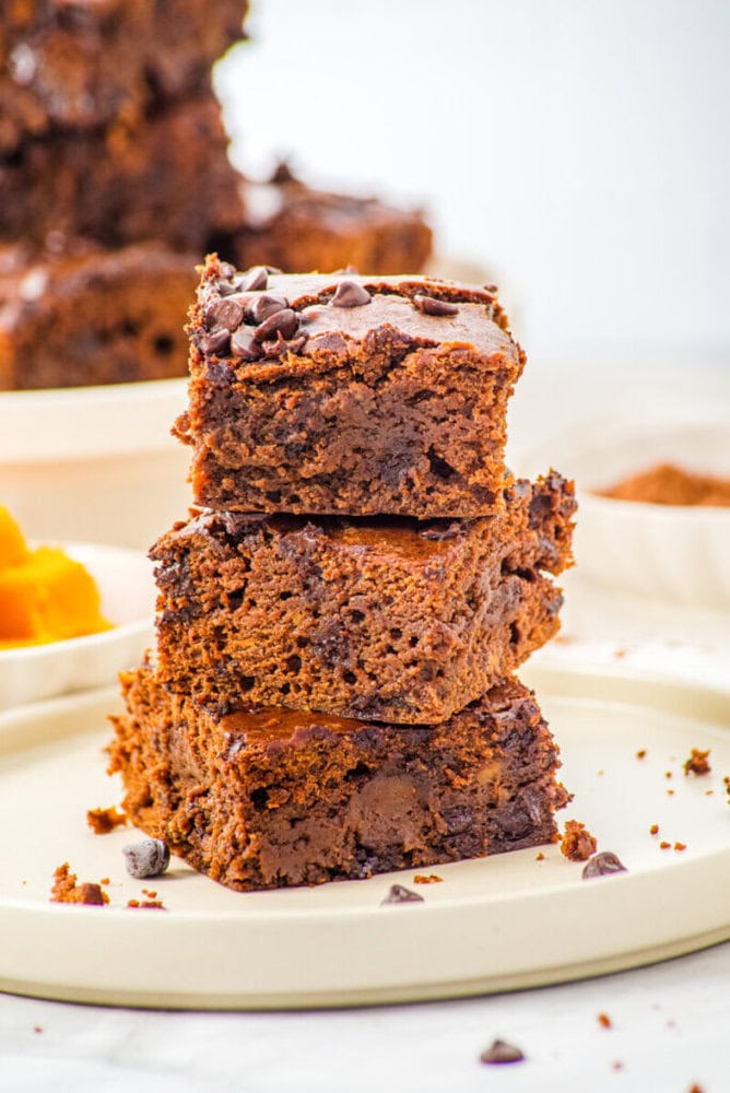 A stack of three chocolate brownies with chocolate chips sits on a round beige plate, with more brownies blurred in the background.