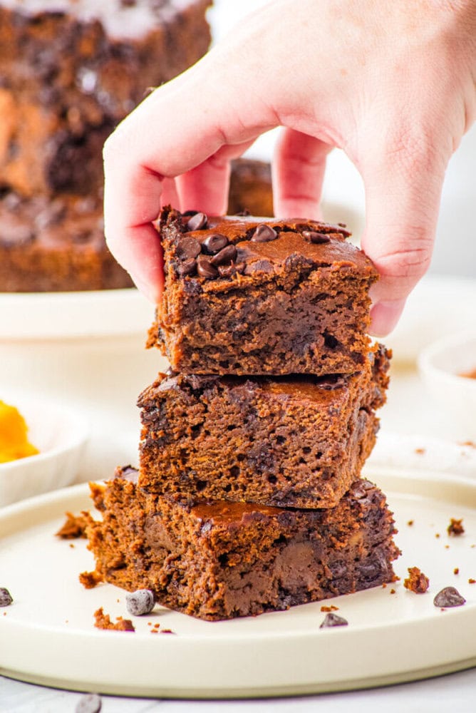A hand placing a brownie square on top of a stack of two brownies, all topped with chocolate chips, on a plate.