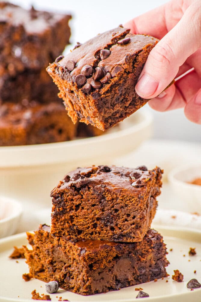 A hand lifts a chocolate chip brownie from a stack of two brownies on a plate, with more brownies and crumbs visible in the background.