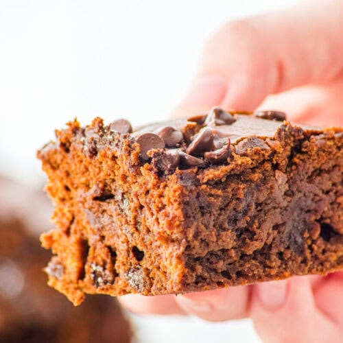 A hand holds a thick, moist chocolate brownie topped with chocolate chips against a blurred background.