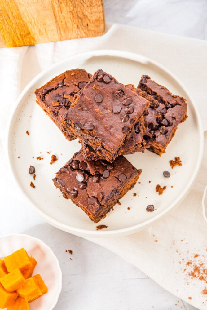 A plate with four chocolate chip brownies stacked on a white surface, with a small bowl of orange cubes nearby.