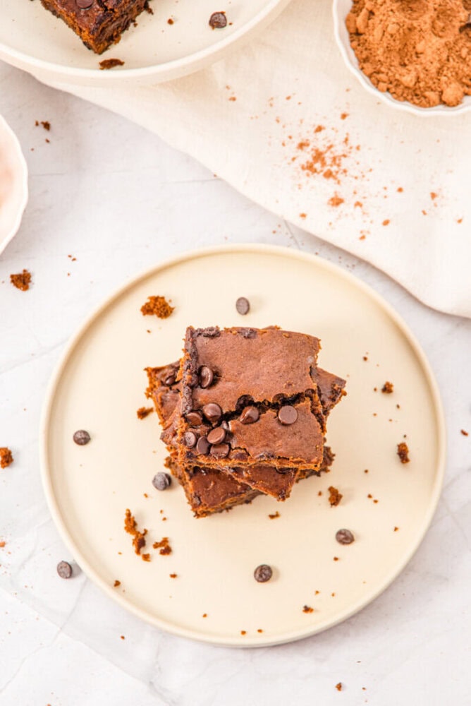 A stack of three chocolate chip brownies on a beige plate, with brownie crumbs and a few chocolate chips scattered around.