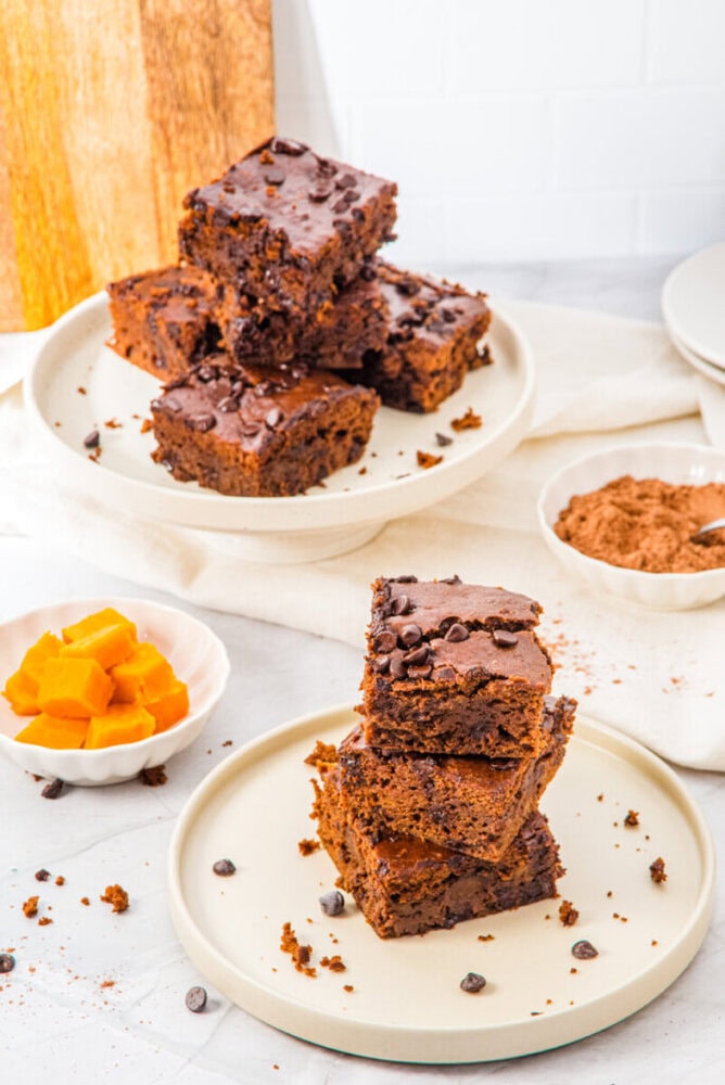 Plates of stacked chocolate brownies with chocolate chips on a white countertop, surrounded by cocoa powder and a bowl of orange cubed ingredients.