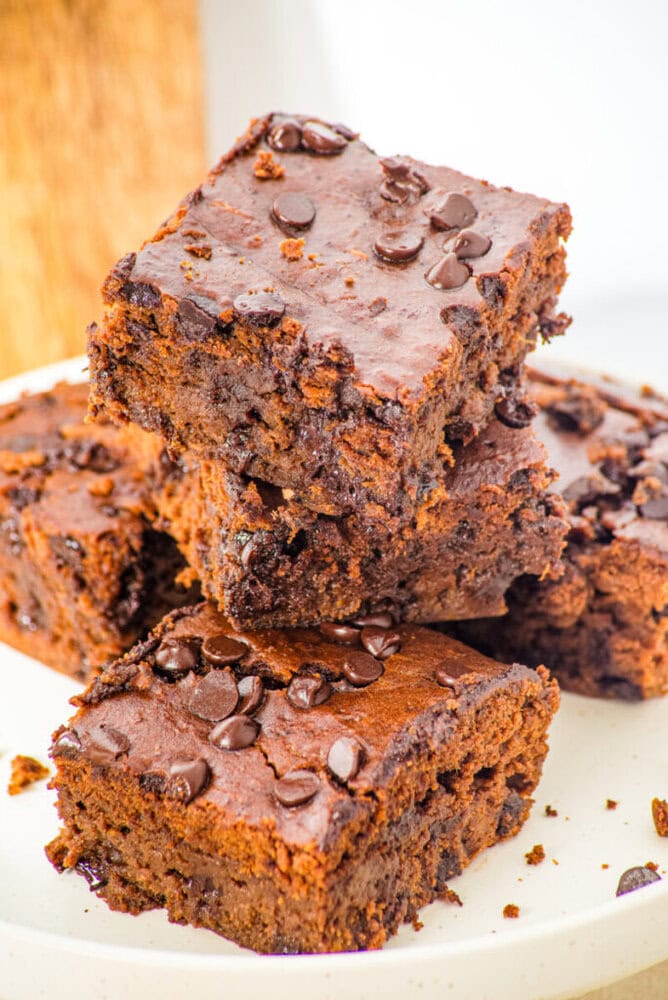 A stack of three chocolate brownies topped with chocolate chips on a white plate, with more brownies visible underneath.