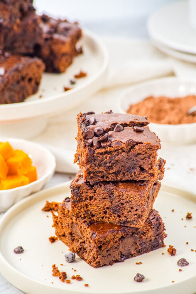A stack of three chocolate chip brownies is displayed on a plate, with more brownies and ingredients visible in the background.