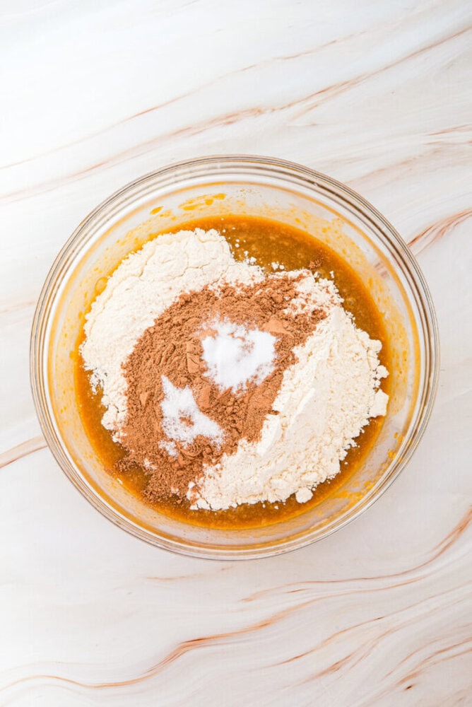 A glass bowl containing flour, cocoa powder, and sugar on top of a brown liquid mixture, set on a marble surface.