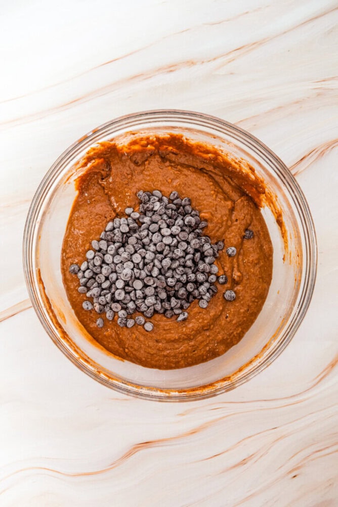A glass bowl filled with chocolate batter topped with a pile of chocolate chips sits on a marble surface.