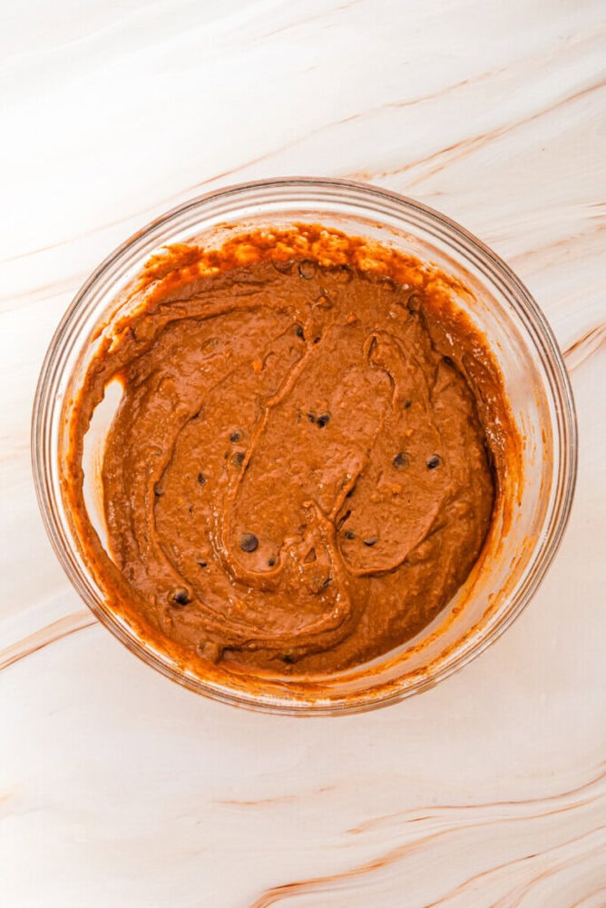 A glass bowl filled with chocolate chip brownie batter sits on a light marble surface.