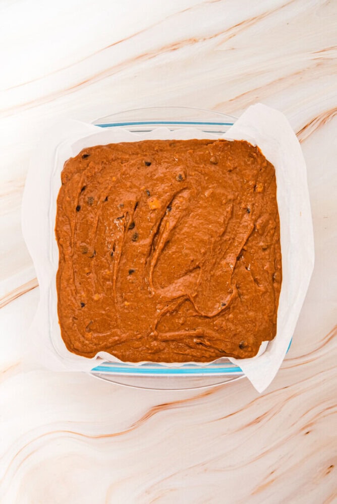 A square glass dish lined with parchment paper, filled with unbaked chocolate batter containing visible chocolate chips, sits on a light marble surface.