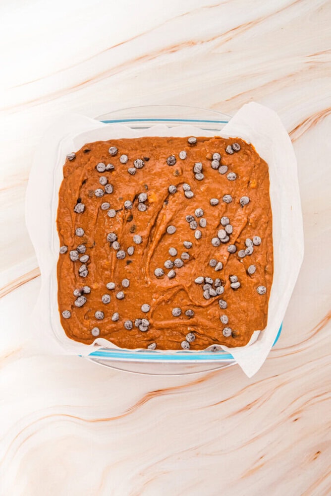 A square glass baking dish lined with parchment paper, filled with chocolate chip-studded batter, sits on a light marble surface.
