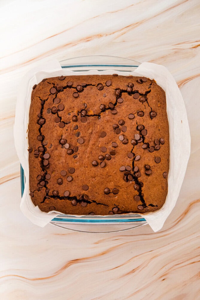 A square chocolate chip cake with cracks on top, baked in a glass dish lined with parchment paper, sits on a marble surface.