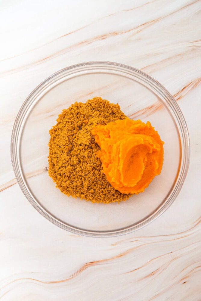 A glass bowl containing a pile of brown sugar and a scoop of pumpkin puree sits on a light marble surface.