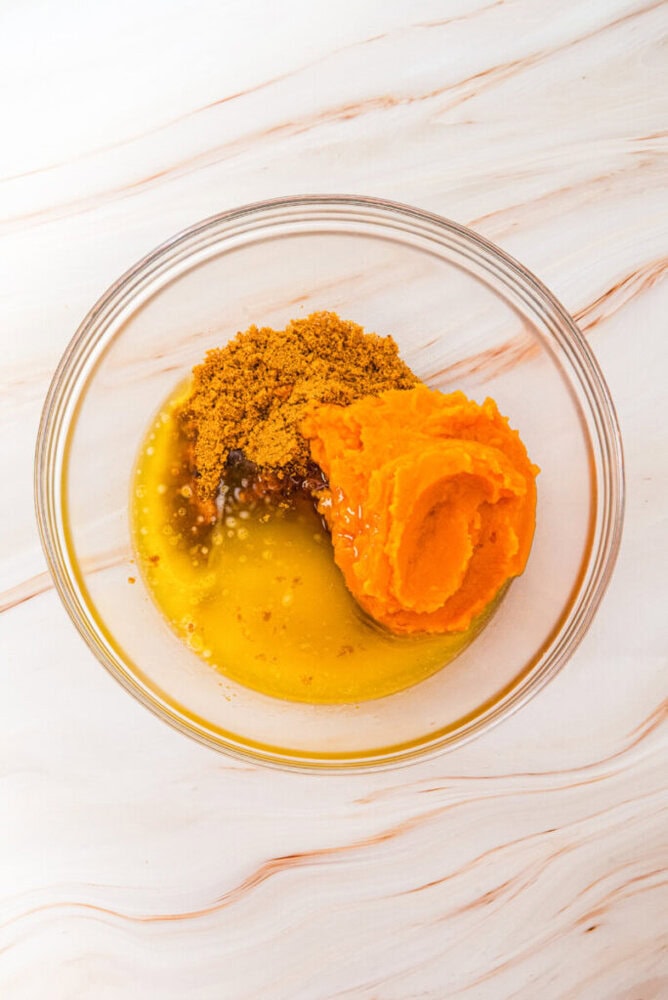 A glass bowl containing pumpkin puree, brown sugar, melted butter, and liquid ingredients on a marble surface.