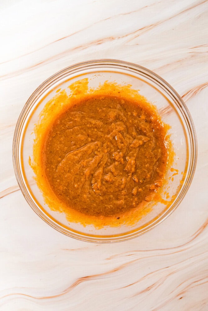 A clear glass bowl containing a thick, orange-brown sauce or marinade sits on a light marble surface.