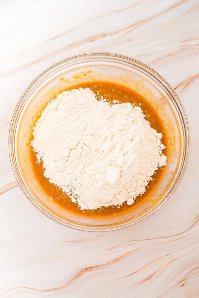 A glass bowl with flour on top of a brown liquid mixture, sitting on a light marble surface, ready to be mixed.