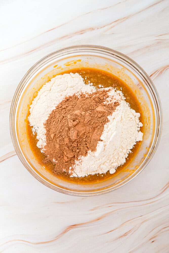 A glass bowl containing flour, cocoa powder, and a brown liquid mixture sits on a white marbled surface, ready to be mixed for baking.